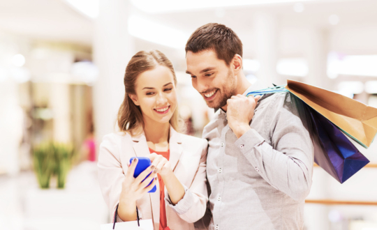 sale, consumerism, technology and people concept - happy young couple with shopping bags and smartphone talking in mall