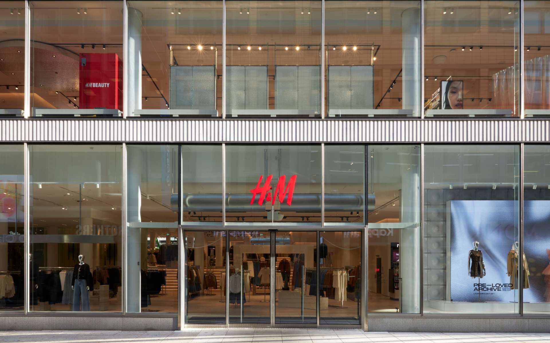 Widok zewnętrzny nowoczesnego sklepu H&M z przeszkloną fasadą. Na środku wejście z logo H&M w czerwonym kolorze. W środku widoczne są ubrania, manekiny i ekspozycje, a po prawej stronie witryna z napisem „Pre-Loved Archive” i dwoma manekinami w ubraniach.